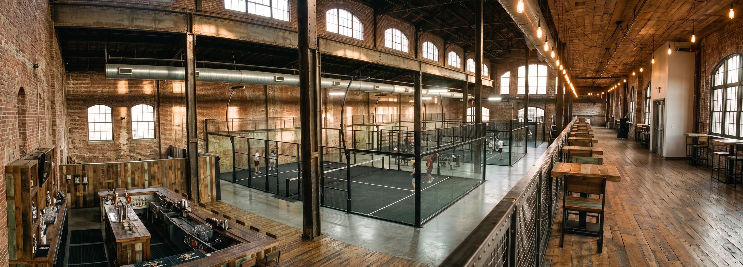 Cancha de pádel Panoramic Crest de Orion XSport en primer plano dentro de un club cubierto de estética industrial, con más canchas al fondo. Fabricación directa de fábrica.