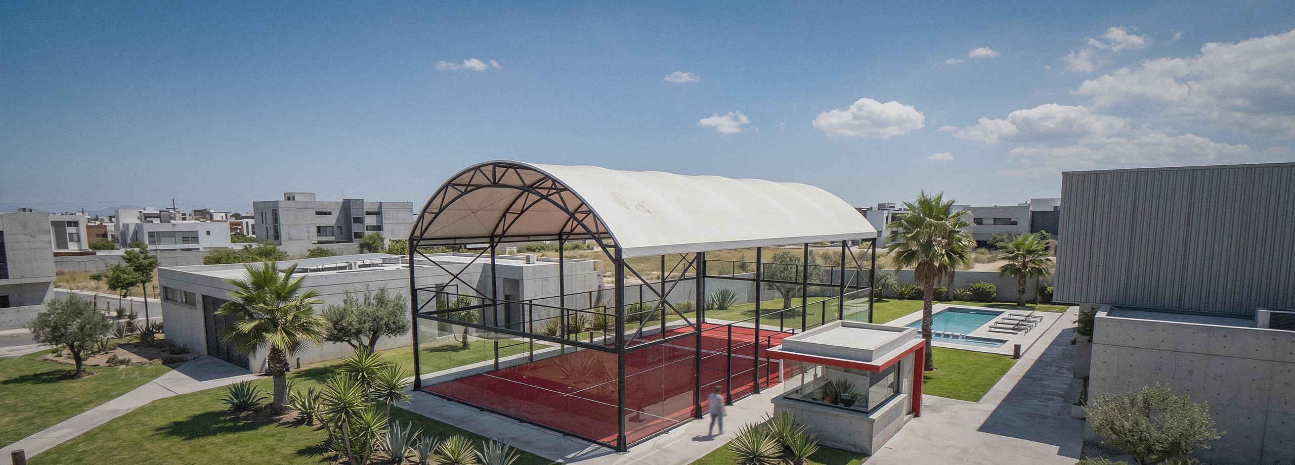 Cancha de pádel Shelter de Orion XSport con su cubierta de arco, en un club deportivo contemporáneo, con césped rojo, caseta de recepción acristalada con banda roja, paisajismo de palmeras y agaves, y piscina junto al acceso. Fabricación directa de fábrica.